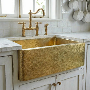 Handmade hammered brass apron sink installed in vintage white kitchen cabinet under window