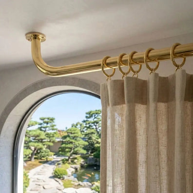 Ceiling-mounted unlacquered brass curtain rod with rings and linen drapes above an arched window