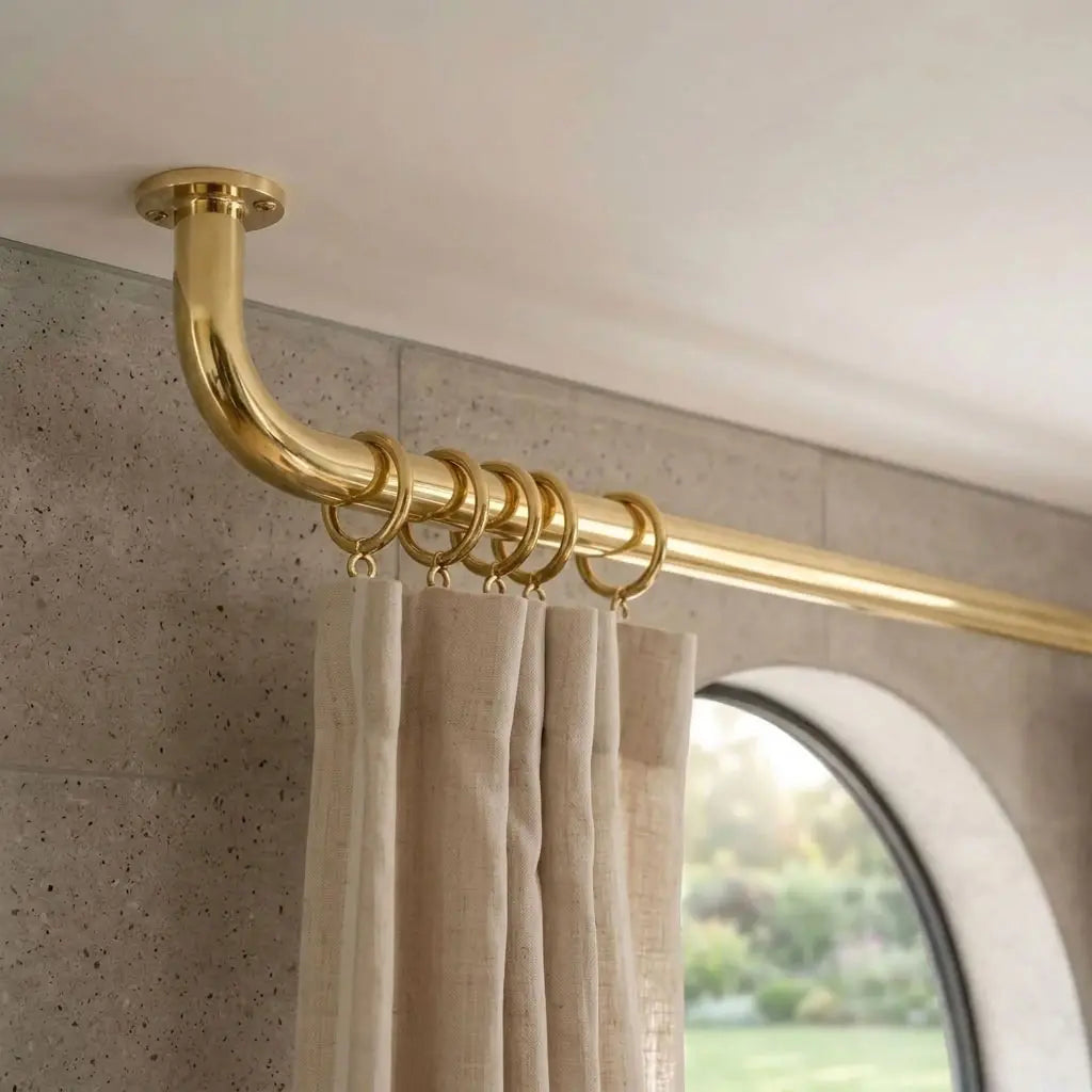 Ceiling-mounted unlacquered brass curtain rod with curved return bracket and matching rings