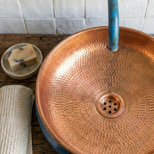 Copper sink with a blue faucet on a wooden surface, next to a bar of soap and a towel.