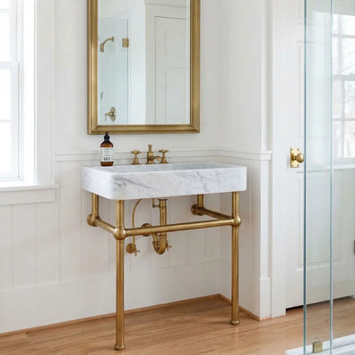 Unlacquered brass console sink base installed beneath a marble sink in a bright bathroom