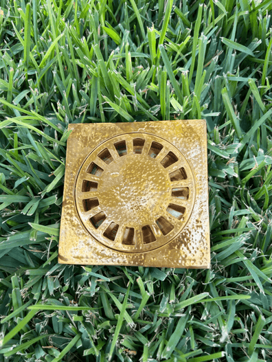 Hammered brass square shower drain with removable cover displayed on grass