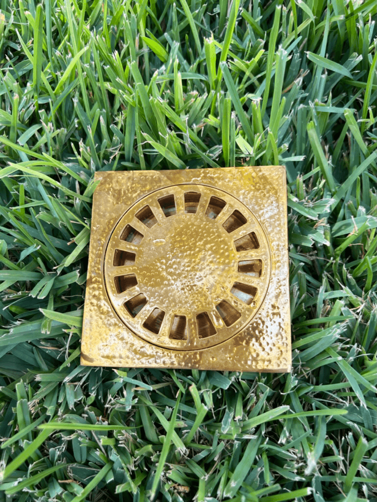 Hammered brass square shower drain with removable cover displayed on grass