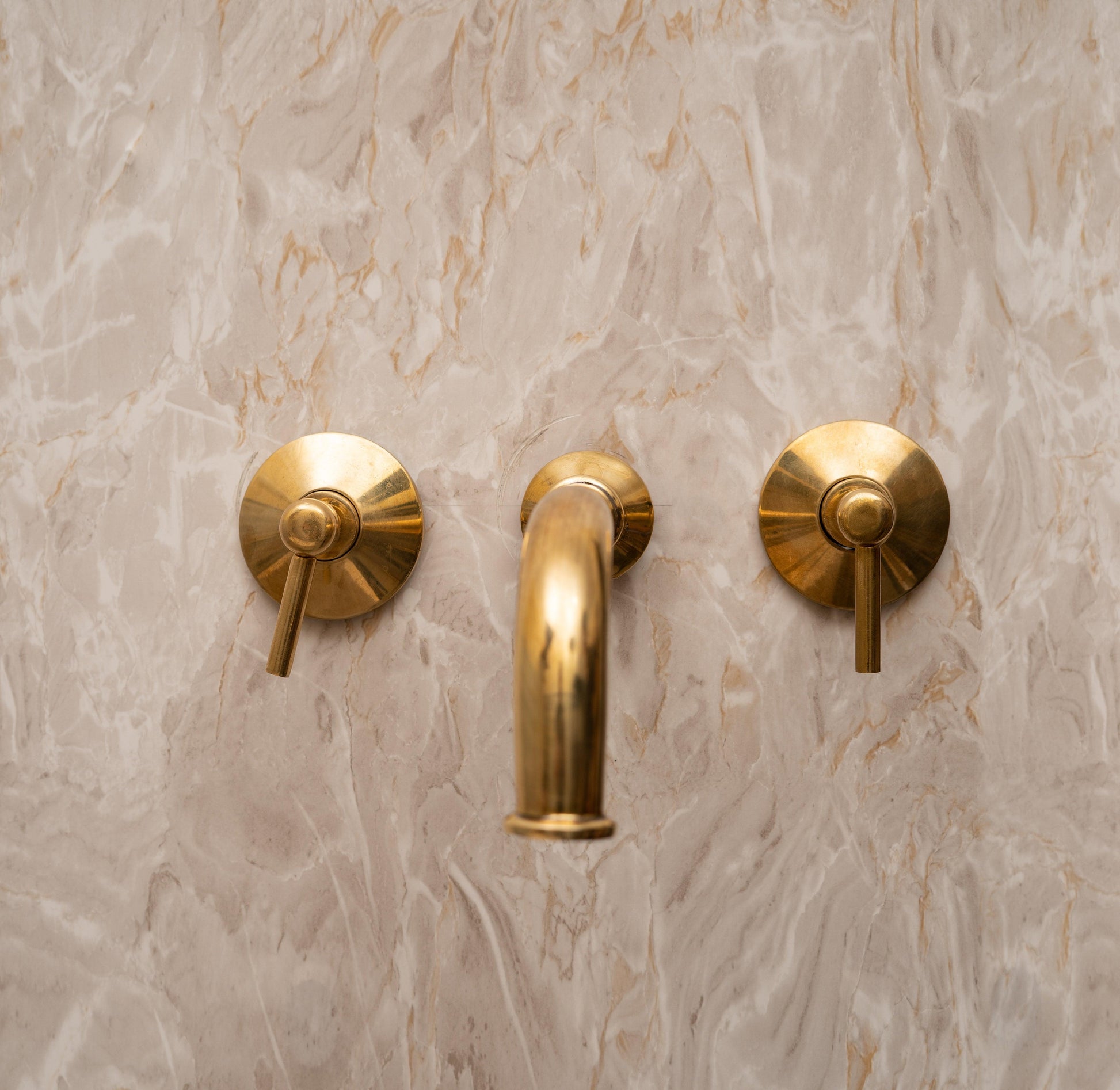Unlacquered brass wall-mount bathroom faucet with lever handles on a marble backsplash