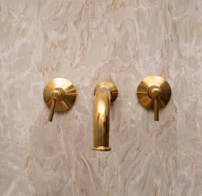 Unlacquered brass wall-mount bathroom faucet with lever handles on a marble backsplash