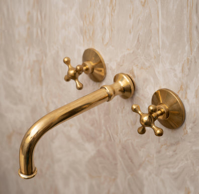 Angled view of an unlacquered brass wall-mounted bathroom faucet with cross handles