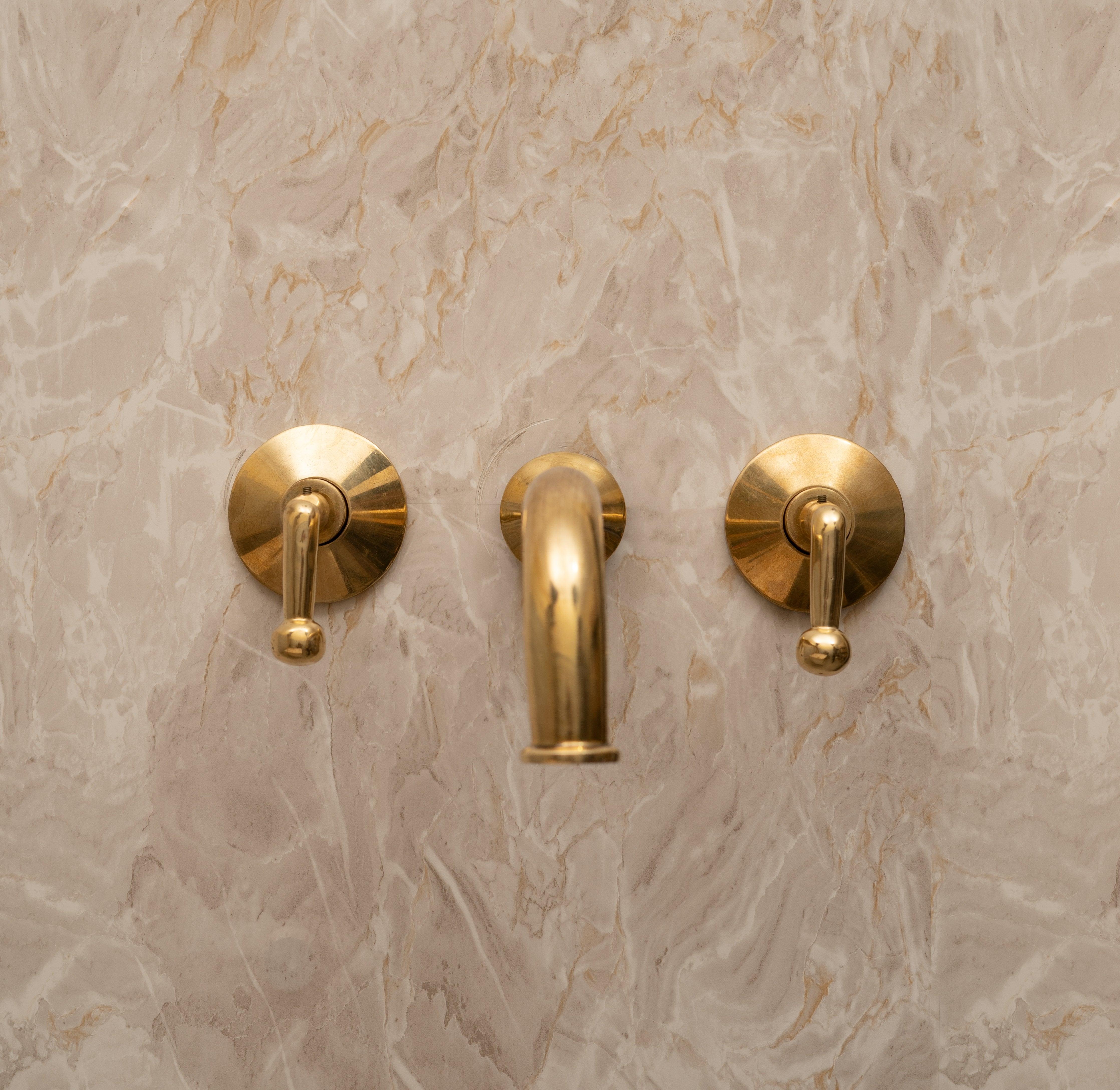 Unlacquered brass wall-mounted bathroom faucet with lever handles installed on marble