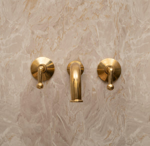Unlacquered brass wall-mounted bathroom faucet with lever handles installed on marble