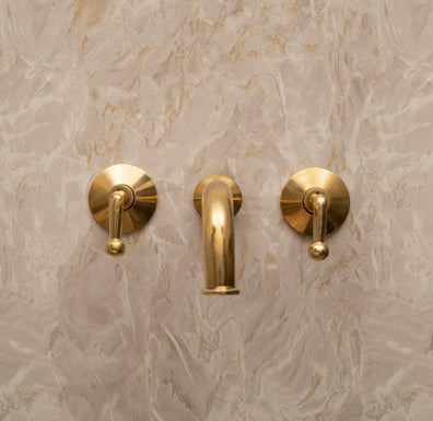 Unlacquered brass wall-mounted bathroom faucet with lever handles installed on marble