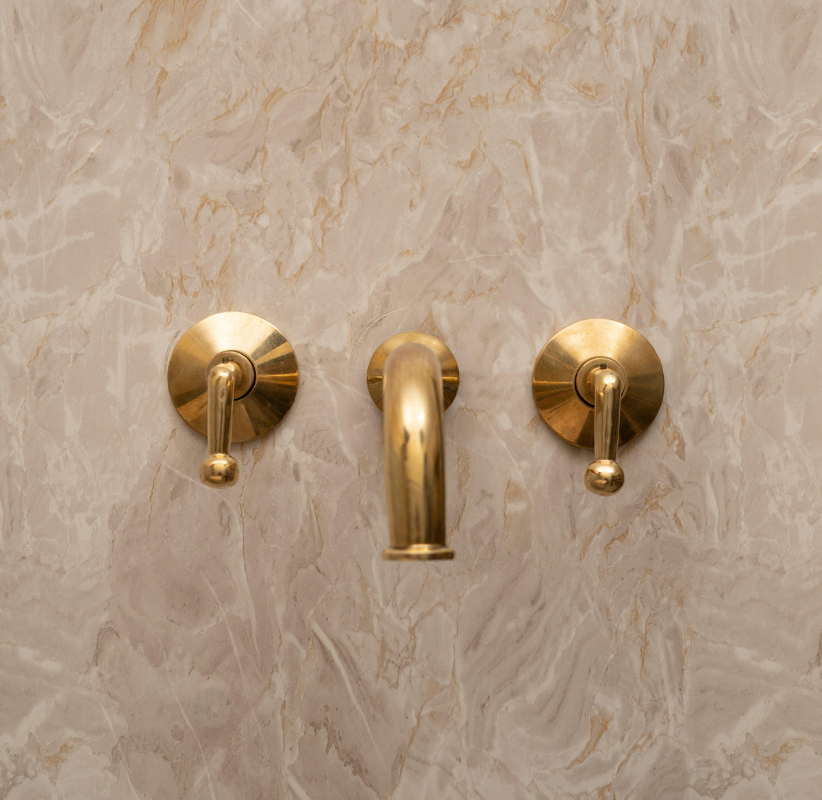 Unlacquered brass wall-mounted bathroom faucet with lever handles installed on marble