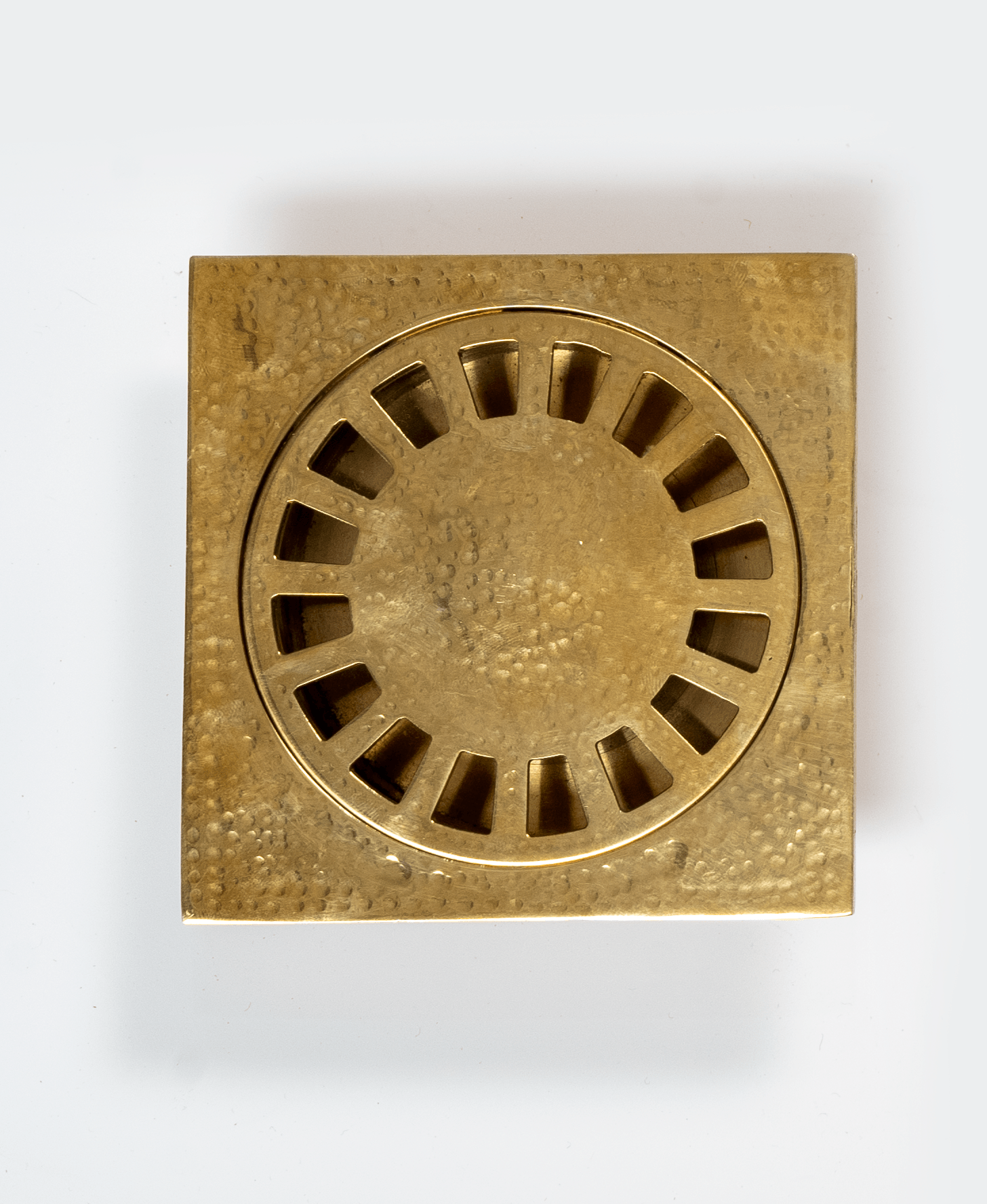 Close-up of an unlacquered hammered brass square floor drain with slotted round grate