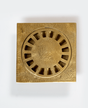 Close-up of an unlacquered hammered brass square floor drain with slotted round grate